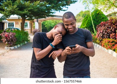 Two Young Black Men Walking And Holding Each Other Laughing Together While Viewing Content On A Mobile Phone. Gay Couple Laughing Together While Using A Phone Together