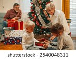 Two young boys are excited to be opening presents on christmas morning, with their grandmother watching and grandfather in the background