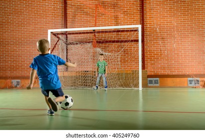 Two young boys playing soccer on an indoor court with one standing in the goalposts as the second prepares to kick for goal - Powered by Shutterstock