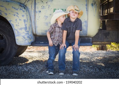 Two Young Boys Wearing Cowboy Hats Leaning Against an Antique Truck in a Rustic Country Setting. 