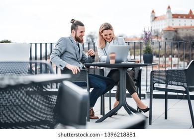 Two Young Business People With Laptop Sitting On A Terrace Outside Office, Working.