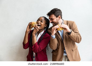 Two Young Business People Or Smart Casual White Man And African American Woman Holding And Eating Fat Tasty American Hamburger Or Burger While They Making Funny Faces And Goofing. Happy People Eating 