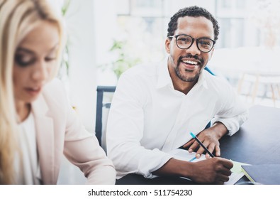 Two Young Business People Working Together In A Modern Office.Black Man Wearing Glasses, Looking At The Camera And Smiling.Businesswoman Discussing With Colleague New Project.Horizontal,blurred