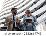 Two young business professionals in sleek corporate attire converse confidently while walking outside their office building