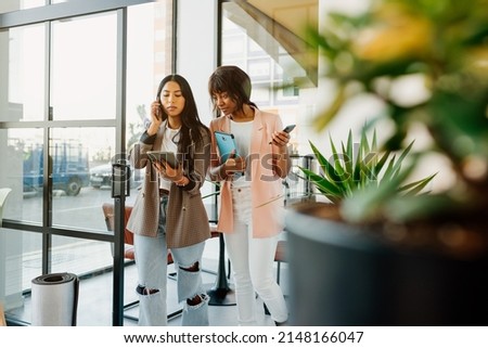 Two young business women of different races working in the office. Latin woman talks on the phone as she walks through the office with her co-worker.