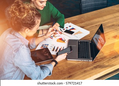 Two Young Business Women Sitting At Table In Front Of Laptop.On Table Is Tablet Computer And Paper Charts.First Woman Uses Smartphone,other Takes Notes In Notebook.Online Education. Instagram Filter.