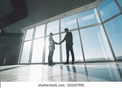 Two young businessmen are shaking hands with each other standing against panoramic windows. - Shutterstock ID 555557905