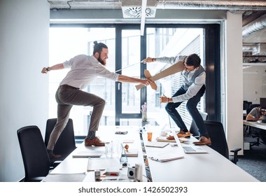 Two young businessmen with swords in an office, having fun. A competition concept.