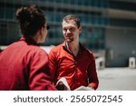 Two young businesspeople engage in a friendly outdoor meeting, wearing casual red attire. The background shows an urban office environment.