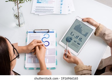 Two young businesswomen analyzing and discussing graph, charts and diagrams while one of them pointing at tablet screen at meeting