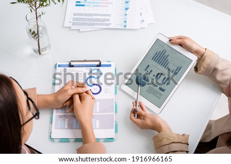 Two young businesswomen analyzing and discussing graph, charts and diagrams while one of them pointing at tablet screen at meeting