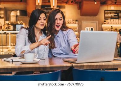 Two young businesswomen, bloggers are sitting in cafe at table and using laptop. Girls with joyful surprise show hand on computer screen, girls look at monitor in amazement, delightedly, joyfully.