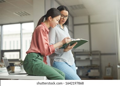 Two Young Businesswomen In Casual Clothing Holding Note Pad And Looking At Their Plans For Work Week Together At Office