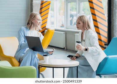 two young businesswomen discussing something, looking at a laptop during a coffee break in the lobby of an industrial office building - Powered by Shutterstock