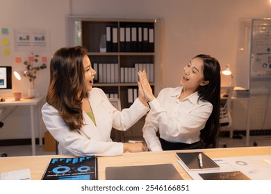 Two young businesswomen are giving each other a high five in celebration of achieving a business target, expressing joy and teamwork in a modern office environment - Powered by Shutterstock