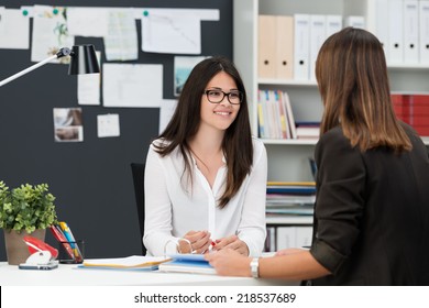 Two young businesswomen having a meeting in the office sitting at a desk having a discussion with focus to a young woman wearing glasses
