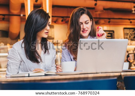 Two young businesswomen sitting in cafe at table and using laptop, working, blogging. Girls are looking at monitor and smiling. Students studying. On desk paper notebook. E-learning, online marketing.