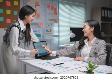 Two young businesswomen smiling and discussing work while analyzing financial graph on tablet in modern office - Powered by Shutterstock