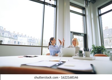 Two young businesswomen smiling and high fiving together while sitting at an office table working on a laptop - Shutterstock ID 1622168146