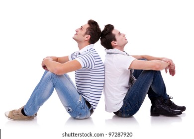 Two Young Casual Men Sitting Back To Back On White Background And Looking At Something On Both Sides Of The Picture