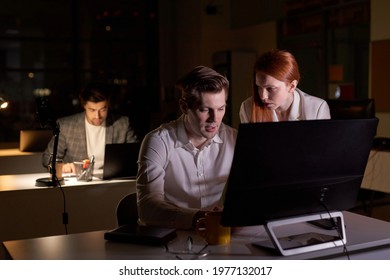 Two Young Caucasian Business People Working Together On Computer, Discussing Project In Office At Late Night. Side View On Redhead Lady And Handsome Guy Colleagues Brainsotrming. In Dark Boardroom