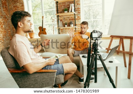 Two young caucasian male bloggers in casual clothes with professional equipment or camera recording video interview at home. Blogging, videoblog, vlogging. Talking while streaming live indoors.