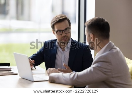 Two young Caucasian male colleagues talking seated at desk in office, discuss teamwork, working together with laptop share ideas, having business conversation during formal meeting in modern workspace