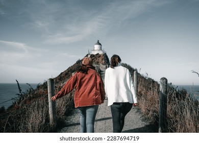 two young caucasian women walking with their backs to the camera towards the pretty summit lighthouse along the dirt road by the fence under a blue sky, nugget point, new zealand - Powered by Shutterstock