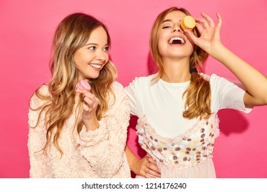 Two young charming  beautiful smiling hipster girls in trendy summer clothes. Women  with colorful macaroons, holding macarons near face. Posing on pink background 