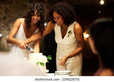 Two young cheerful brides in elegant wedding attire cutting big cake while standing by festive table in front of guests enjoying party - Powered by Shutterstock