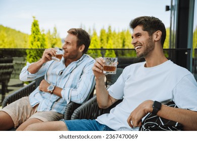 Dos jóvenes y alegres amigos hombres con ropa informal bebiendo whisky mientras se relajan en la terraza en las montañas