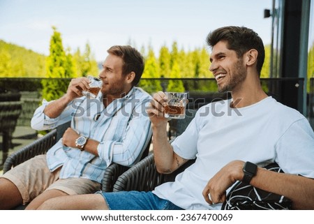 Two young cheerful male friends in casual wear drinking whiskey while relaxing on terrace in mountains