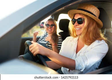 Two young cheerful smiling women in a car on vacation trip to the sea beach. Girl in glasses driving a vehicle from rental on holidays. Girlfriends enjoying summer arrived to ocean shore on holidays.