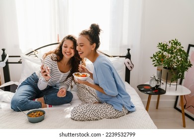 Two young cheerful white girls eating pasta and using mobile phone while sitting on bed together at home - Powered by Shutterstock