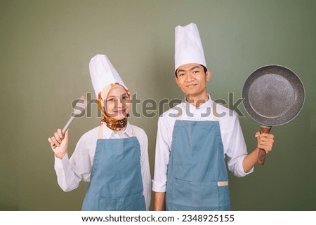 two young chef in cooking clothes holding a frying pan and a spatula with a smiling expression in front of a green background