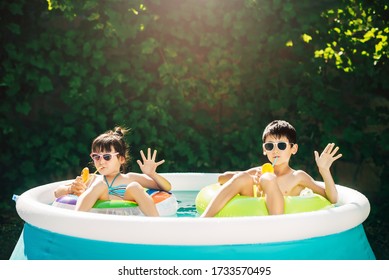 Two Young Children Enjoy Summer Vacation And Drink Orange Juice From Glasses While Swimming On Inflatable Circles In The Home Pool In The Yard. Hello Gesture. Kids Sunbathe.