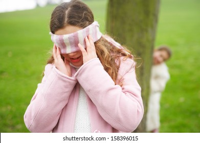 Two young children and family sisters playing hide and seek together in a park with green grass and hiding behind a tree while blindfolded and counting during a cold winter day.