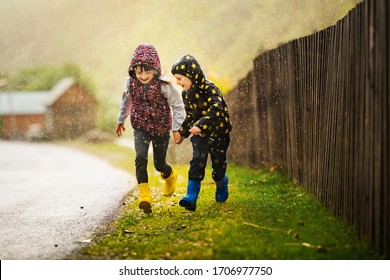 Two young children laughing running in spring or autumn rain. Boy and girl holding hands. Children are wearing hooded jackets and rubber boots. Kids have fun time playing outdoors under summer shower - Powered by Shutterstock