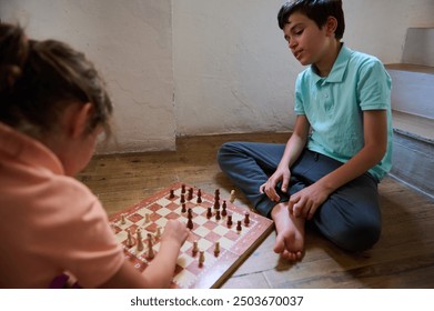 Two young children sit cross-legged on the floor, absorbed in a chess game. Their expressions reflect concentration and problem-solving skills in a relaxed home environment. - Powered by Shutterstock