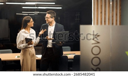 Two young colleagues having conversation using laptop and tablet behind glass wall in board room, creative director and project manager discussing work