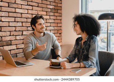 Two young colleagues using laptop while drinking coffee in cafeteria. Coworkers customers couple having conversation, doing freelance job projects online in cafe restaurant - Powered by Shutterstock