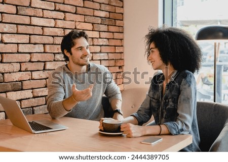 Two young colleagues using laptop while drinking coffee in cafeteria. Coworkers customers couple having conversation, doing freelance job projects online in cafe restaurant