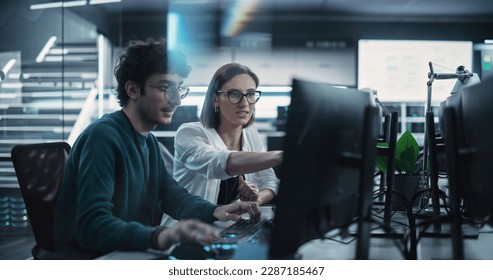 Two Young Colleagues Working on Computers and Talking at a Workplace. Female and Male Software Developers Discussing a Solution for Their Collaborative Artificial Intelligence Project - Powered by Shutterstock