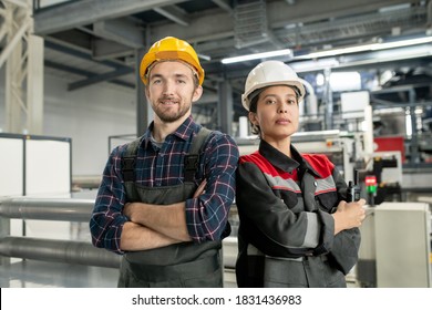 Two young confident workers in helmets and workwear crossing arms by chest while standing against interior or workshop of modern factory