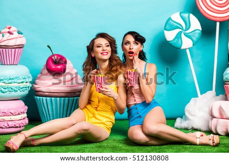 Two young cool lady on turquoise background, sitting on the grass and eating popcorn, laughing, best friends, crazy emotions, Huge candy, lollipops, cotton candy, Cake, fashionable Pin up girl