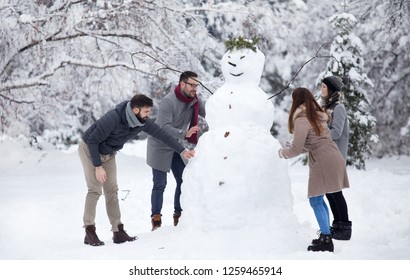 Two young couples having fun by building big snowman in park during winter holidays