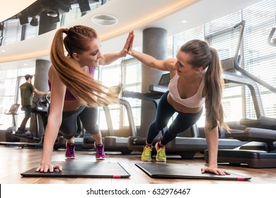 Two young and determined beautiful women giving high five while practicing basic plank exercise on mats during workout in a modern fitness center - Powered by Shutterstock