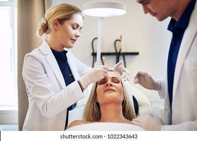 Two young doctors discussing a cosmetic surgery procedure on the face of a mature woman lying on a table in a clinic