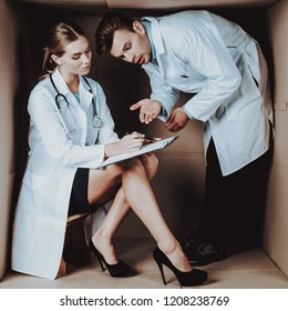 Two Young Doctors In White Coats In Cardboard Box. Young Woman In White Coat. Life In Little Cardboard Box. Uncomfortable Life. Personal Spase Concepts. Medical Equipment. Doctor Witn Tablet.