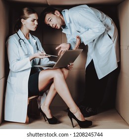 Two Young Doctors In White Coats In Cardboard Box. Young Woman In White Coat. Life In Little Cardboard Box. Uncomfortable Life. Personal Spase Concepts. Medical Equipment. Doctor Witn Laptop.